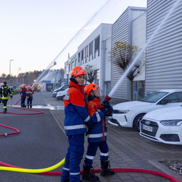 Ein größerer Junge von der jugendfeuerwehr unterstützt einen kleineren Jungen beim abgeben eines Strahls mit dem Schlauch.