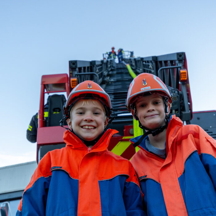 Zwei lächelnde Kinder von der Jugendfeuerwehr stehen vor der Drehleiter. Im Korb der Drehleiter stehen zwei andere Kinder mit einem Löschschlauch.
