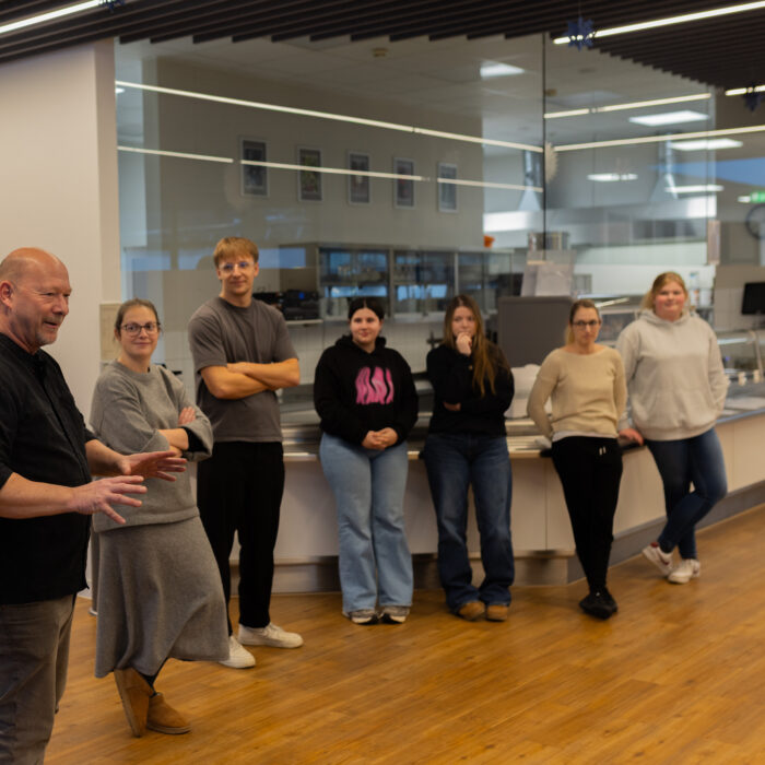 Sechs Späh-Mitarbeiter und ein Koch stehen in einem Halbkreis in der Kantine. Der Koch erklärt den Mitarbeitern den Ablauf für das Backen vom Weihnachtsgebäck.