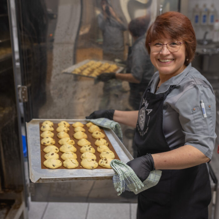 Eine lächelnde Späh-Mitarbeiterin aus der Kantine hat ein Backblech voller Weihnachtsgebäck in ihren Händen. Die Mitarbeiterin trägt Handschuhe, da das Blech vor kurzem aus dem Ofen geholt wurde.