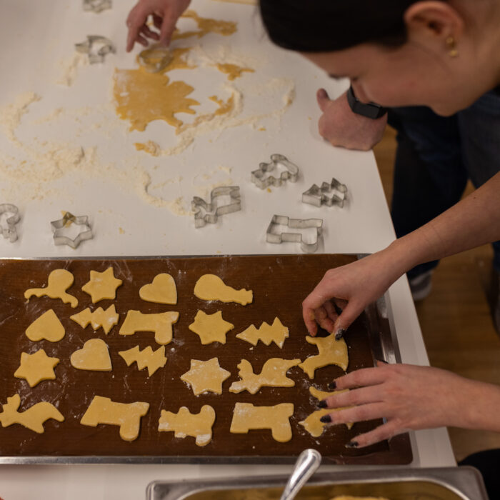 Zwei Späh-Mitarbeiter die Weihnachtsgebäck backen. Der eine Mitarbeiter legt ausgestochene Plätzchen auf das Backblech. Der andere Mitarbeiter sticht gerade weitere Plätzchen aus. Auf dem Tisch sind viele unterschiedliche Formen zum Ausstechen verteilt.