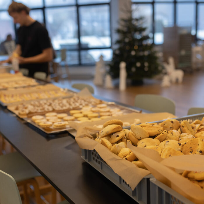 Auf einem langen Tisch stehen viele Bleche mit unterschiedlichen fertigen Weihnachtsgebäcken. Ein Späh-Mitarbeiter steht vor dem Tisch und hält eine Packung Puderzucker.
