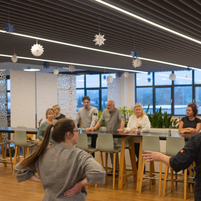 Viele Späh-Mitarbeiter und ein Koch stehen versammelt in der Kantine. Der Koch erklärt den Späh-Mitarbeitern das Vorgehen für das Backen vom Weihnachtsgebäck.