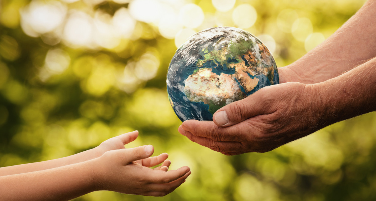 Eine erwachsene Person hält ein kleines Modell der Erde in beiden Händen und übergibt diese einem Kind was beide Hände offen hält.